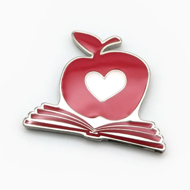 Pin de esmalte de una manzana roja con un corazón calado, sobre un libro abierto.