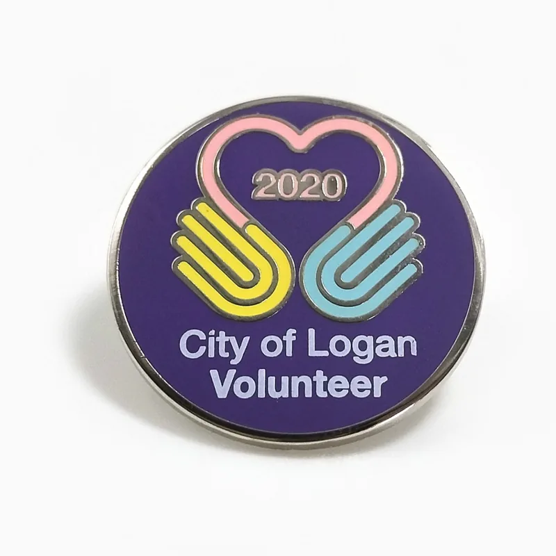 Pin redondo de esmalte duro para voluntariado con dos manos que forman un corazón sobre esmalte morado y el año 2020