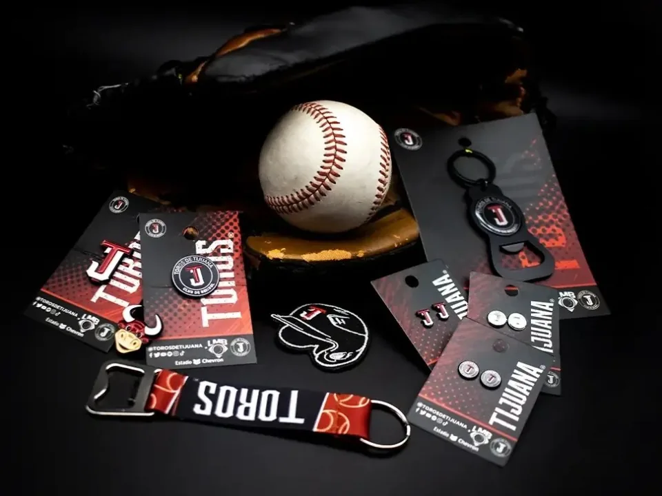 Pins personalizados, llaveros, aretes y un abridor de botellas en tarjetas de presentación con marca, junto a una pelota y un guante de béisbol.