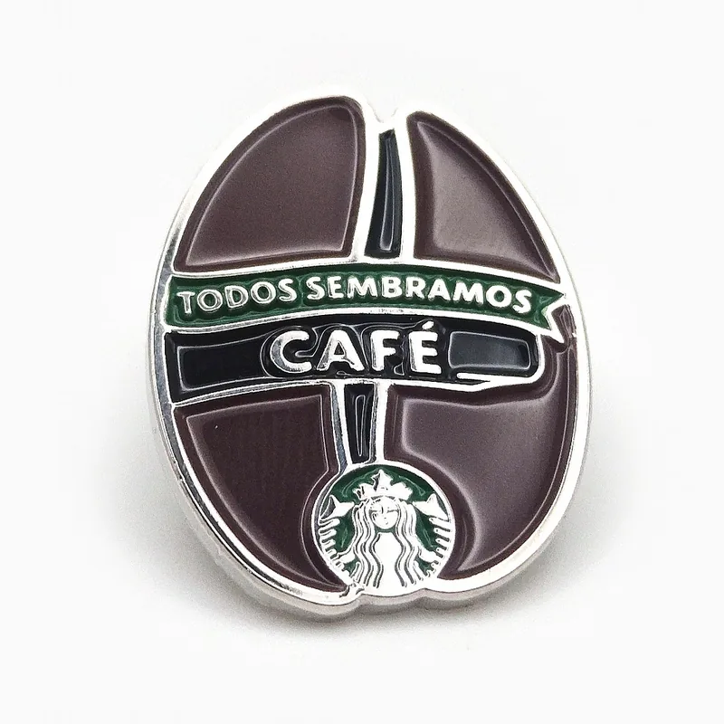 Pin de esmalte con forma de grano de café y una cinta plateada que dice “Café”, hecho para una cadena de cafeterías.