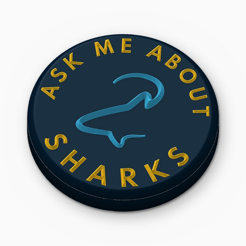 Pin redondo de PVC suave azul marino con el texto Ask me about sharks en amarillo en relieve y la silueta de un tiburón azul